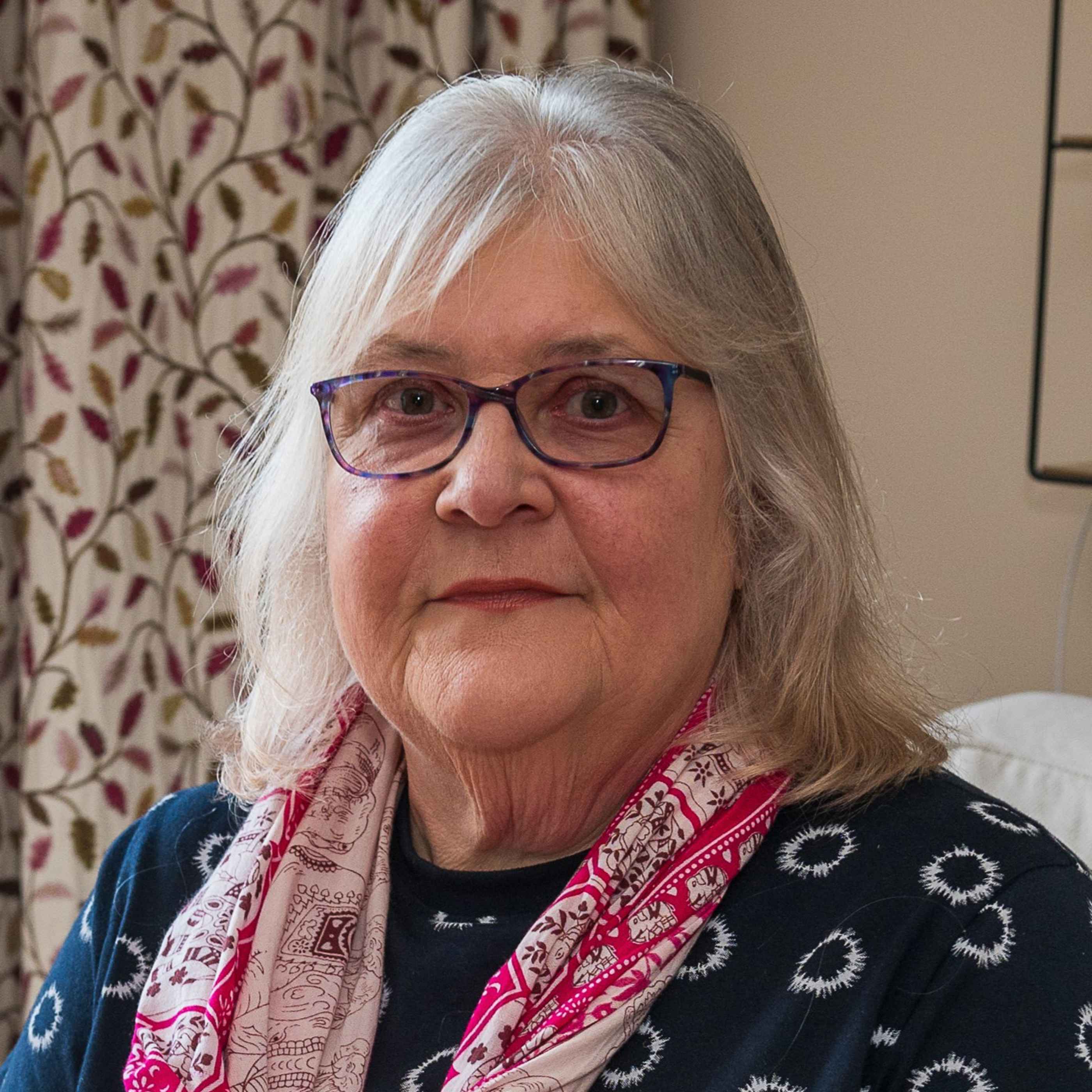 Headshot of Lynne. She is indoors, wearing a navy top with white circular patterns and a red scarf.